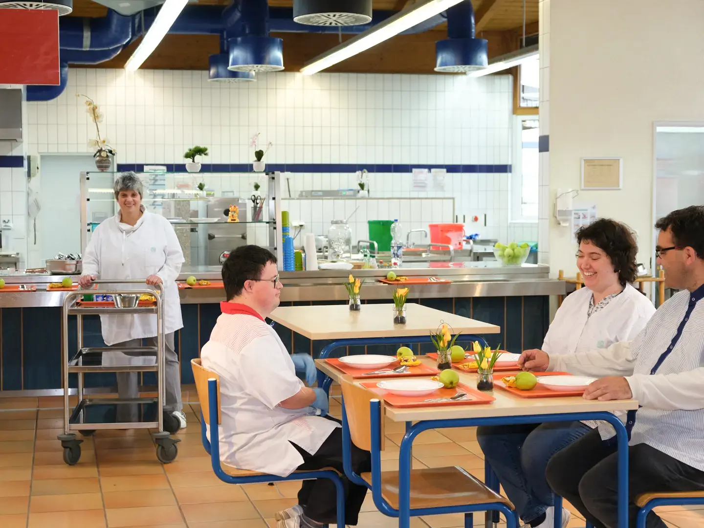 Betriebskantine mit Mitarbeiterin bei der Essensausgabe und Gästen am Tisch