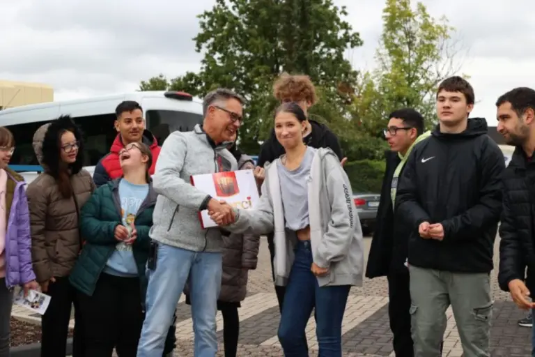 eine Gruppe Schülerinnen und Schüler mit Mitarbeitenden der REHA-Betriebe Erftland auf dem Werksgelände. Der Ausbildungsleiter schüttelt einer Schülerinn die Hand und überreicht ihre eine sehr große Packung Mercie-Schokolade.