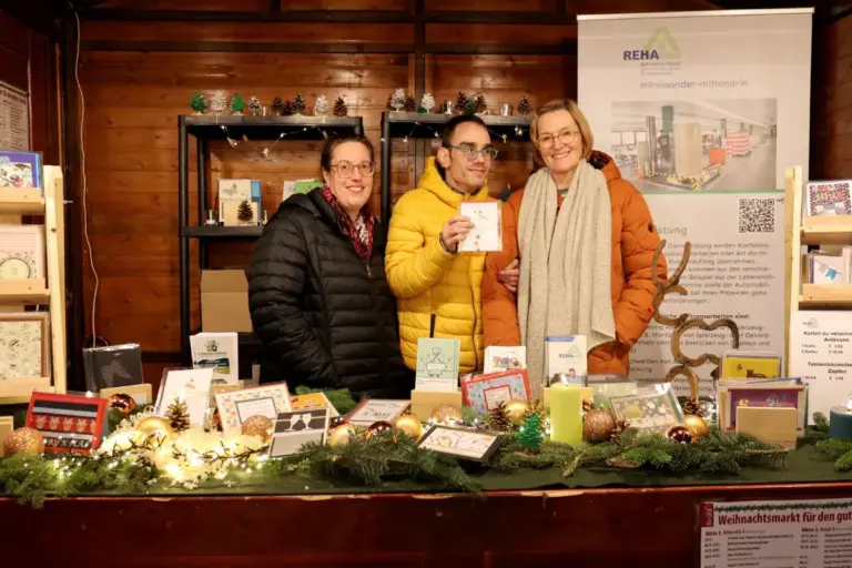 Drei Personen in der festlich geschmücken Weihnachtsmarkt-Bude der REHA-Betriebe Erftland. Der Mann in der Mitte hält eine von den Werkstattbeschäftigten gefertigte Grußkarte in der Hand.