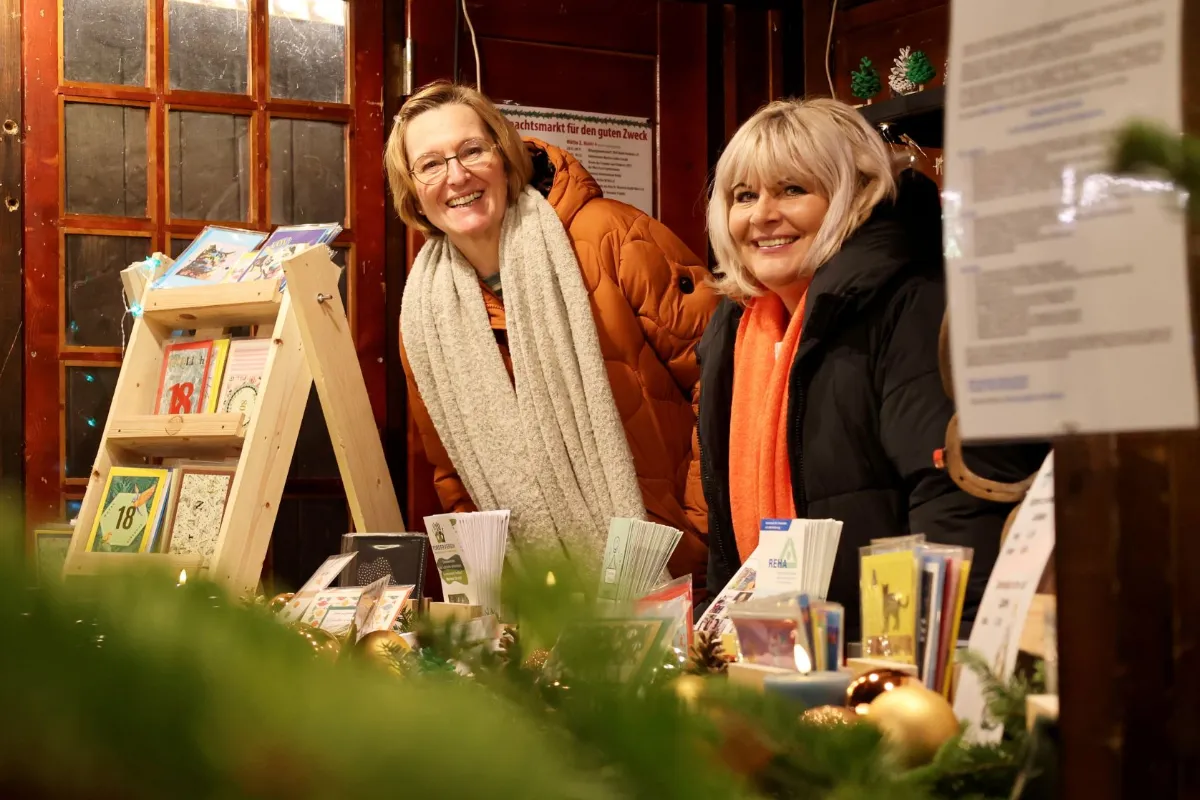 Zwei Damen schauen aus der festlich geschmückten Weihnachtsmarkt-Bude fröhlich in die Kamera