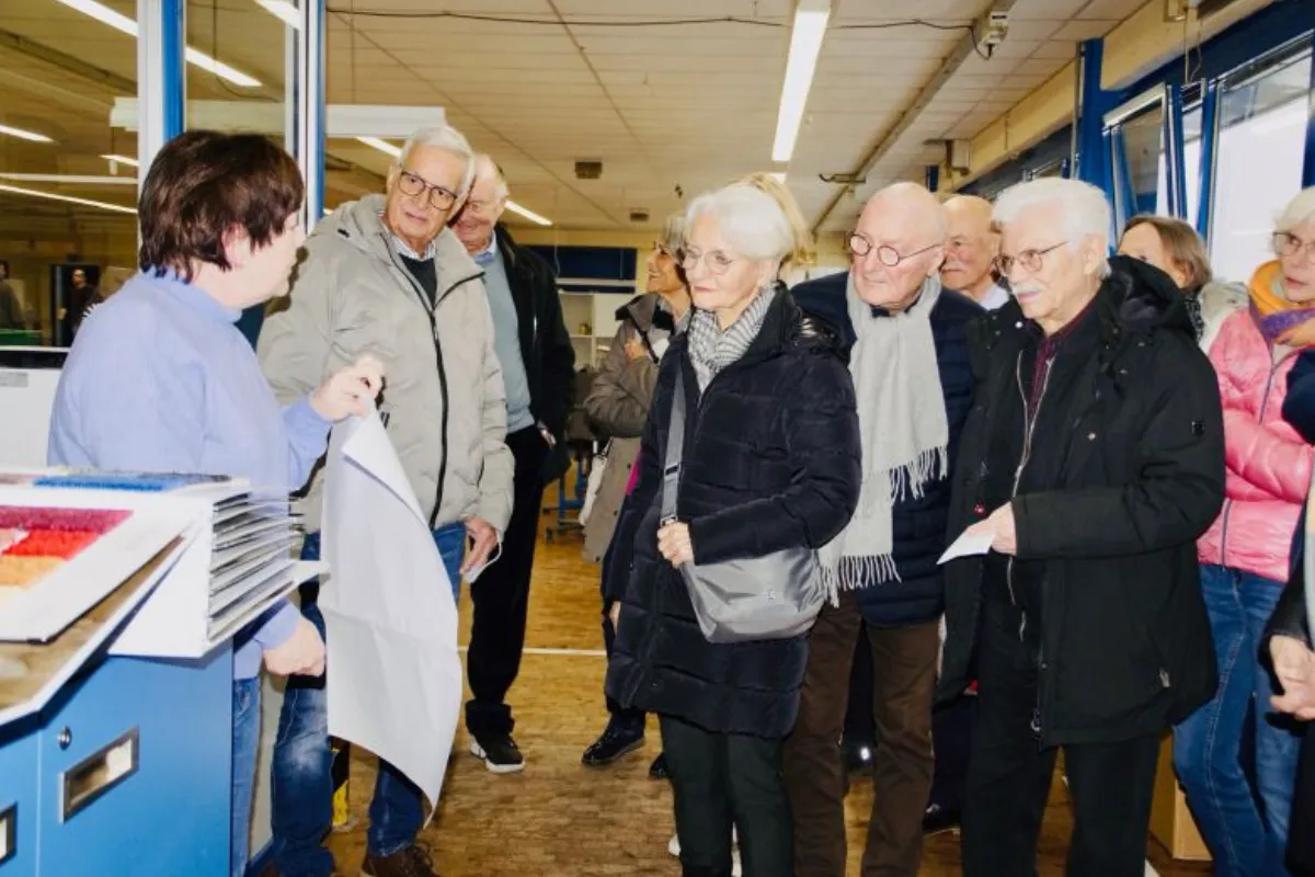 Eine Gruppe von Menschen steht in einer Produktionsumgebung und hört aufmerksam einer Präsentation zu. Eine Person zeigt einige Materialien und erklärt etwas.